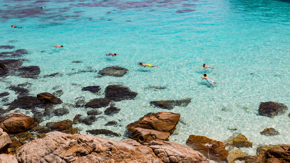 Snorkelers in clear turquoise water beside the rocky shore of Perhentian Besar, Malaysia